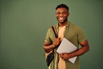 Distance learning. Glad african american guy with backpack and wireless laptop posing over green background. Enthusiastic smiling student engaging in online education with help of modern gadget. - obrazy, fototapety, plakaty