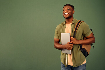Online studying. Handsome afro young guy in casual khaki shirt cheerfully smiling while posing over green background with backpack on shoulder and laptop in hand. Copy space for text and advertising.