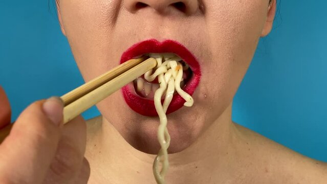 Unrecognizable young adult woman vibrantly colored lips eating noodles using chopsticks, having delicious lunch indoors. Close-up mouth red lipstick savoring pasta or ramen isolated on blue background