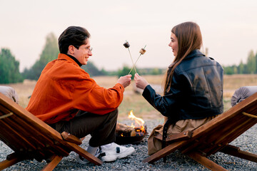 Back shot of young man and woman in love feeding each other toasted marshmallow and admiring scenery of nature - obrazy, fototapety, plakaty