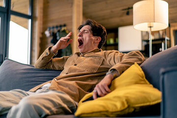 A young guy yawns sitting on the sofa with  remote control in his hand watching TV at home