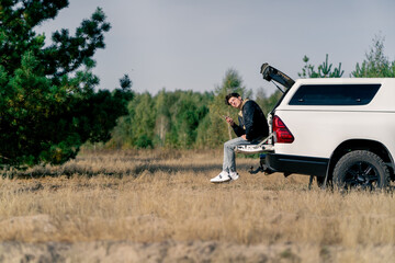 The car is standing on the road in the forest-steppe A young guy sits in the open trunk with phone and gets connection