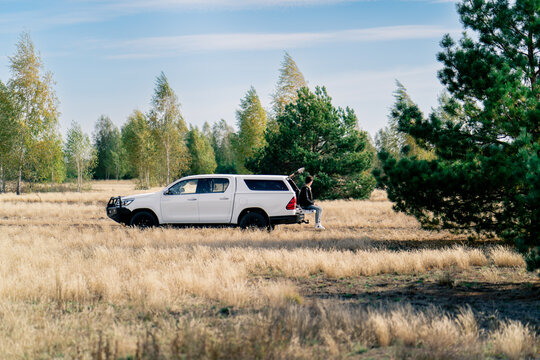 The car is standing on the road in the forest-steppe A young guy sits in the open trunk with phone and gets connection - Powered by Adobe