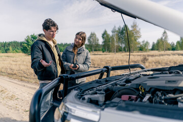 A guy driver helps a girl fix her car in field and forest during a trip by opening the hood