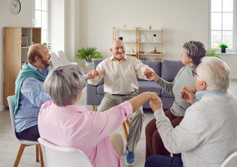 Positive seniors sitting in circle and holding hands during group therapy in nursing home. Smiling mature men and women sitting together on chairs in living room during support group session.