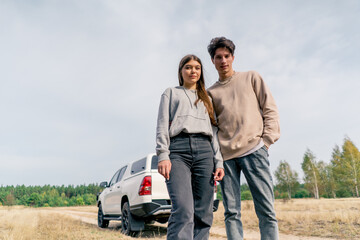 Beautiful friends a guy and a girl stand against the background of car in the forest steppe smiling and holding hands