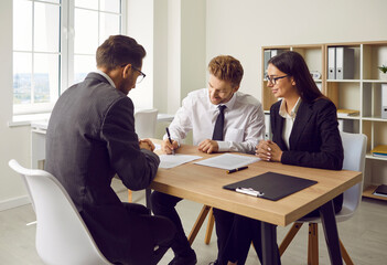 Portrait of a group of three coworkers and company employees sitting at the desk reaching agreement on meeting. Confident young business man make a deal signing a contract at the office.