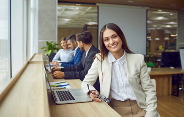 Portrait of young happy smiling business woman sitting in modern open plan office at same desk with coworkers with laptop and looking cheerful at camera. Female company employee working on workplace.