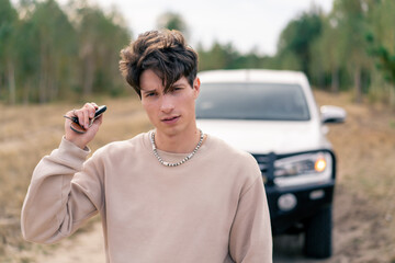 A young successful guy stands with car keys on the road in forest against background of his SUV