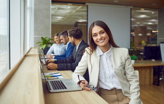 Portrait Of Young Happy Smiling Business Woman Sitting In Modern Open Plan Office At Same Desk With Coworkers With Laptop And Looking Cheerful At Camera. Female Company Employee Working On Workplace.