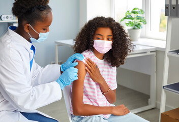 A female doctor of African-American appearance disinfects the shoulder area for injection to the patient. An African-American teenage girl is preparing to get vaccinated against coronavirus or flu.