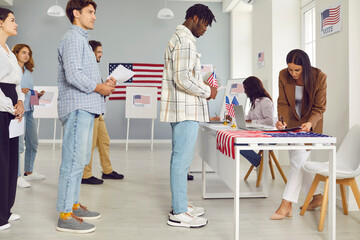 Diverse US voters at polling place. Multiracial multiethnic Caucasian and African American people standing in line waiting for their turn to come up to table of poll worker. USA elections concept - obrazy, fototapety, plakaty