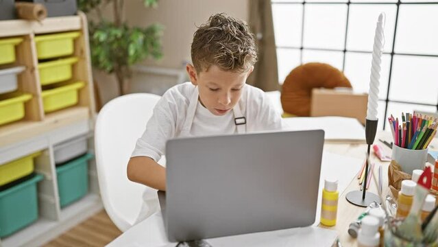 Adorable blond artist boy seriously concentrating on laptop at art studio, sitting in a cute apron, immersed in online painting lesson