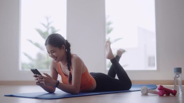 Asian young woman in workout attire is lying on her stomach, using her mobile phone to chat with friends or engage in social networking apps while on a yoga mat in her home gym.