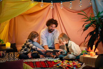 Obraz na płótnie Canvas A good looking dad with curly hair is spending his time with his two young boys bonding and reading a fun book together as they are all in a very big handmade blanket fort indoors