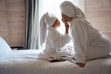Little cute girl in bathrobe massaging mother's face with special device
