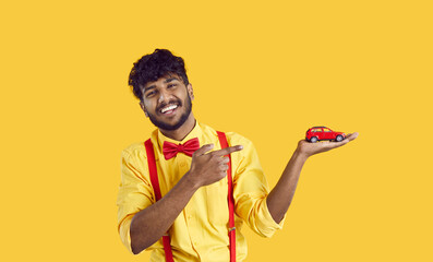 Cheerful Indian guy points his finger at small toy car in his hand, isolated on orange background....