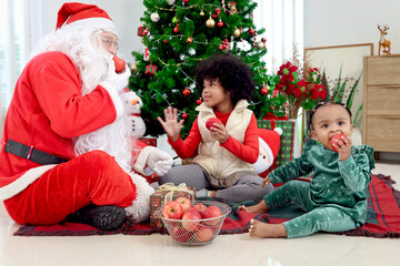 Obraz na płótnie Canvas African child sister and Santa Claus celebrate winter holiday together, two cute girl kid and Sata eating apples under beautiful Christmas tree surrounded by present gift boxes, happy childhood family