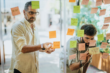 Diverse business team brainstorming, collaborating, and writing ideas on sticky notes on a glass...