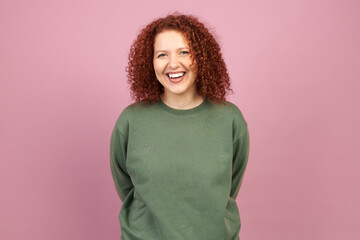 Cheerful redhead caucasian millennial woman with curly hair standing against pink background with...