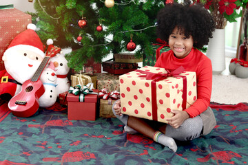 Portrait of happy smiling African kid girl with curly hair holding gift box present while sitting under decorative Christmas tree. Child celebrates winter holiday. merry Christmas, happy childhood