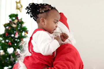 Little African cute girl is carried by Santa Claus in red dress costume with decorative beautiful Christmas tree blurred background. Happy baby kid and Santa celebrate winter holiday. Merry Christmas