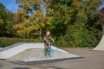 Boy on scooter makes a trick and enjoying his riding in the skate park at cloudy spring day. Young man doing trick on the kick scooter in the park. Full body little boy on the kick scooter on sunny
