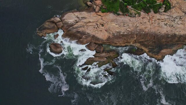 Drone Footage of Ipanema Beach in Rio De Janeiro in Brazil. The video is a 90 degree down view of the pedra do arpoador with waves crashing on it.