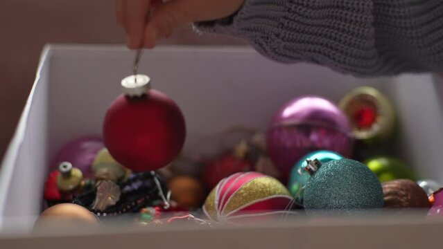 Close-up hands of unrecognizable child girl takes out festive Christmas tree ball from box with various toys. Preparation for holidays, decorates Christmas tree for New Year at home, slow motion.