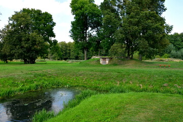 A view of a channel or lake overgrown with moss and vines, with forests and moors on both sides combined with small concrete bridges, walkways, orchards, and a single manor in the middle