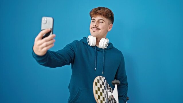 Young hispanic man holding skate wearing headphones make selfie by smartphone over isolated blue background