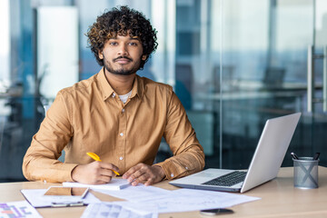 Portrait of young serious thinking businessman inside office behind paperwork, financier investor looking focused at camera, workplace, with papers, reports and contracts.