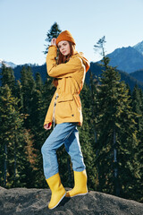 Young woman with red hair full-length hiker in yellow raincoat and cap runs and walks on a mountain trip in the fall and hiking in the mountains at sunset freedom