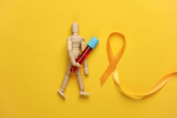 Yellow ribbon and wooden puppet with blood test tube, childhood cancer awareness day symbol