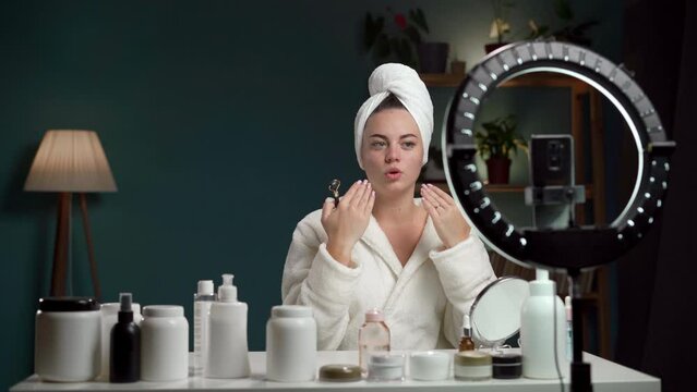 Female blogger in towel and bathrobe is showing face roller for beautiful skin while recording video and giving advices for her beauty blog, in the evening at home