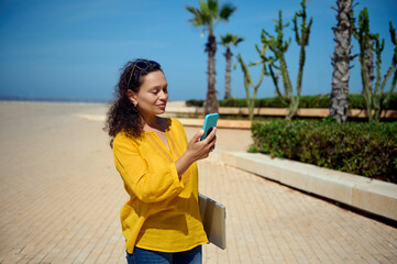 Confident Latin American woman in casual clothes, strolling the street with laptop and smart mobile phone in her hands