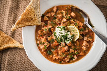 Ost Deutsche DDR GDR Soljanka Eintopf Suppe mit Jagdwurst, Saure Gurken, Letscho, Schmand, Zitrone, Toast und Petersilie mit Löffel, Salzstreuer und braun Stoff Hintergrund