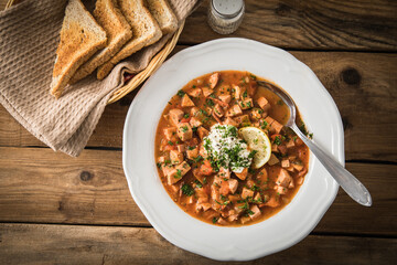 Ost Deutsche DDR GDR Soljanka Eintopf Suppe mit Jagdwurst, Saure Gurken, Letscho, Schmand, Zitrone, Toast und Petersilie auf Holzbrett mit Brotkorb, Salzstreuer auf Holz Hintergrund