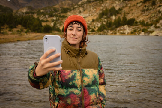 Woman in nature wearing winter hat, hiking in middle of mountains with lake in background looking at happy camera, making photos with the mobile phone making a call