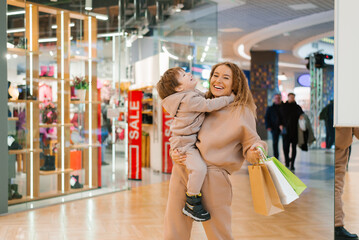 Smiling mother carries her little happy son in her arms, holds shopping bags in her hands and walks...