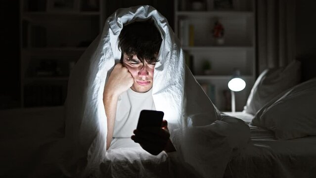 Young hispanic teenager seriously engrossed in texting on his smartphone, lying on a comfy bed under a snug blanket, in the dim light of his cozy bedroom