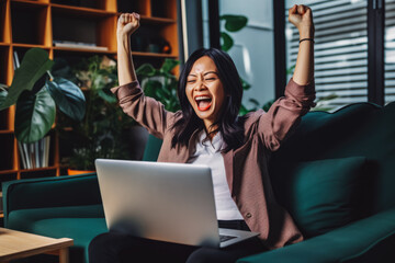 Beautiful middle age asian woman smiling and rejoicing after success. Happy woman celebrating business success on sofa in living room with computer.
