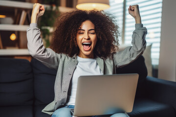 Beautiful young multiethnic woman smiling and rejoices after success. Happy woman celebrating business success on sofa in living room with computer. - obrazy, fototapety, plakaty