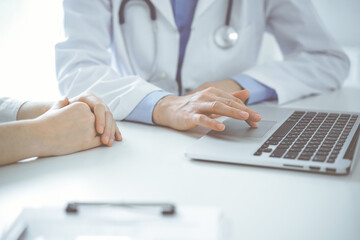 Doctor and patient sitting near each other at the desk in clinic. The focus is on female physician's hands using laptop computer, close up. Medicine concept