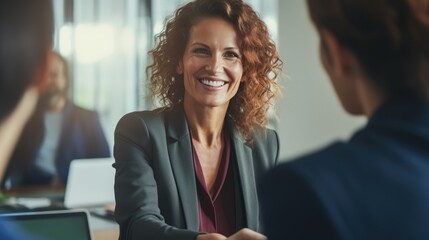 An upbeat, middle-aged businesswoman who is a manager is seen shaking hands during an office meeting. She's greeting a smiling female HR professional conducting a job interview