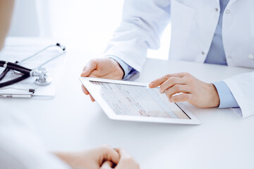 Doctor and patient sitting opposite each other at the desk in clinic. The focus is on female physician's hands pointing into tablet computer touchpad, close up. Medicine concept