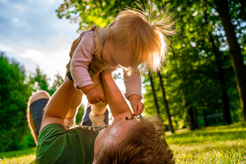 Father plays with a cute blonde baby toddler in the park at sunset, dad lifts the baby up in his arms. The concept of a happy childhood, a strong family. Sun rays with selective focus