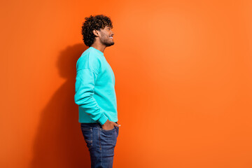 Side shot of young arabian guy wearing stylish bright sweatshirt put hands in jeans look novelty isolated on orange color background