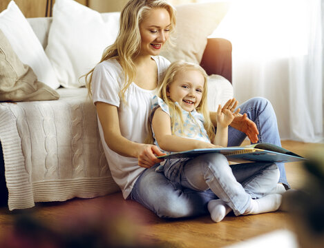 Happy family. Blonde young mother reading a book to her cute daughter while sitting at wooden floor in sunny room. Motherhood concept