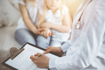 Doctor and patient. Pediatrician using clipboard while examining little girl with her mother at home. Sick and unhappy child at medical exam